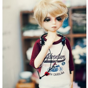 May include: A doll with short blonde hair wearing a white and red baseball shirt with the text "Adventure Motorcycle Club Since 1983 Est 1963 North Carolina" and blue jeans.