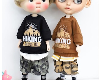 May include: Two dolls with large eyes and stylized hair, one blonde and one dark-haired. Both wear long-sleeve sweatshirts with a mountain graphic and the text HIKING MODE ON, layered over white shirts, camouflage cargo shorts, striped socks, and sneakers.