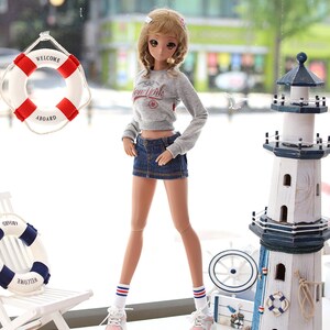 May include: A doll wearing a grey sweater with "New York" printed on it, a denim skirt, white socks with red and blue stripes, and sneakers. The doll is standing on a white surface with a white chair, a lighthouse, and a life preserver.