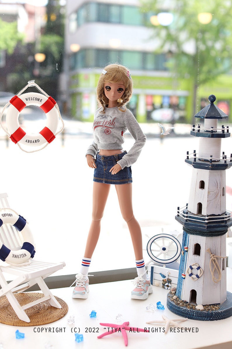 May include: A doll wearing a grey sweater with "New York" printed on it, a denim skirt, white socks with red and blue stripes, and sneakers. The doll is standing on a white surface with a white chair, a lighthouse, and a life preserver.