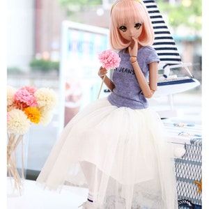 K&ouml;nnte beinhalten: Eine Puppe mit rosa Haaren, die ein hellblaues T-Shirt mit dem Wort "Jeans" tr&auml;gt, einen wei&szlig;en T&uuml;llrock und blaue Turnschuhe. Die Puppe h&auml;lt eine rosa Blume und sitzt auf einer wei&szlig;en Oberfl&auml;che mit einem Segelboot im Hintergrund.