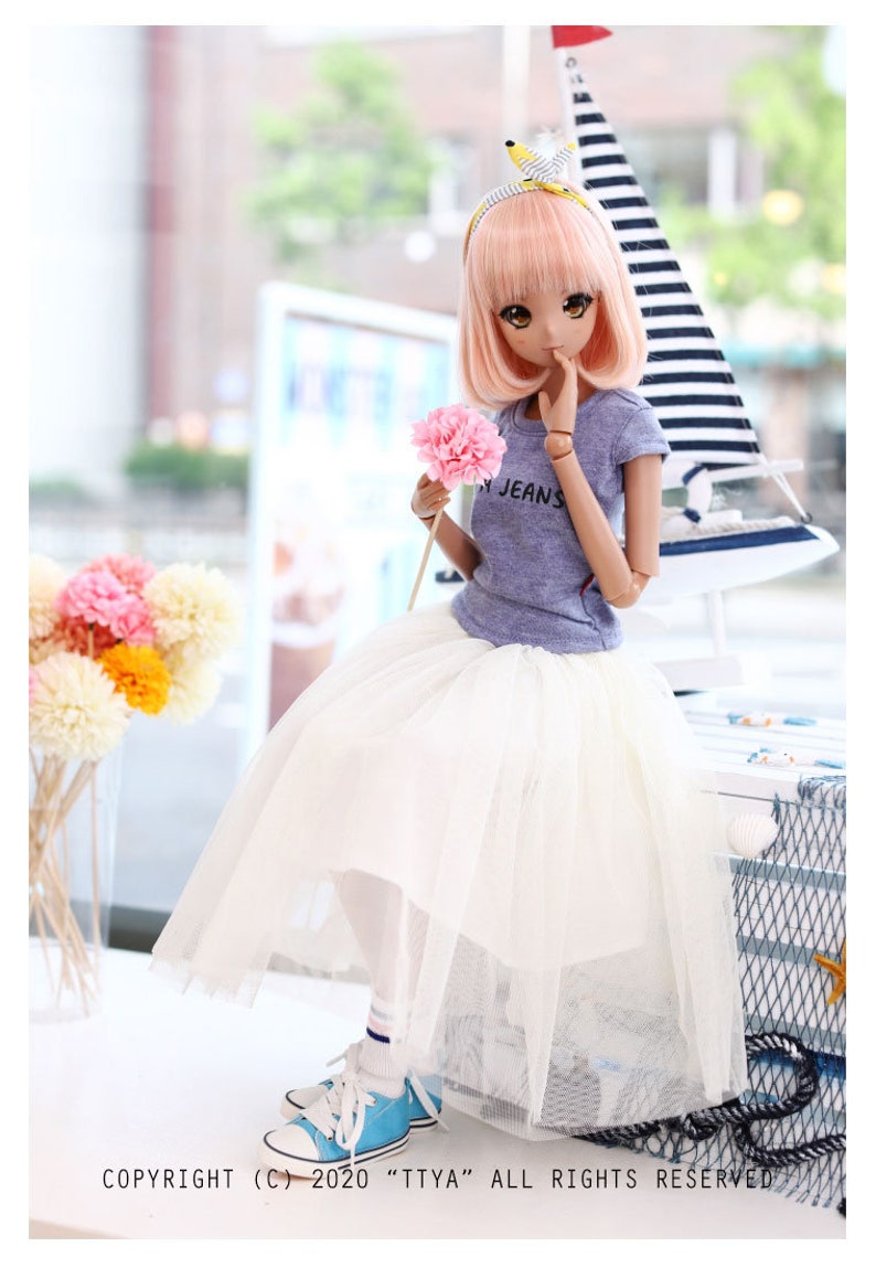 K&ouml;nnte beinhalten: Eine Puppe mit rosa Haaren, die ein hellblaues T-Shirt mit dem Wort "Jeans" tr&auml;gt, einen wei&szlig;en T&uuml;llrock und blaue Turnschuhe. Die Puppe h&auml;lt eine rosa Blume und sitzt auf einer wei&szlig;en Oberfl&auml;che mit einem Segelboot im Hintergrund.