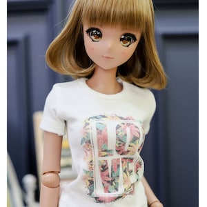 May include: A doll wearing a white t-shirt with a floral print and the word "LOVE" in a box. The doll has brown hair and brown eyes.
