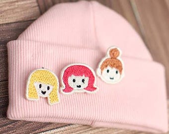 May include: A light pink ribbed knit beanie featuring three embroidered patches of cartoon faces with different hair colors, including yellow, bright pink, and light brown, arranged in a horizontal row on the folded cuff of the hat.