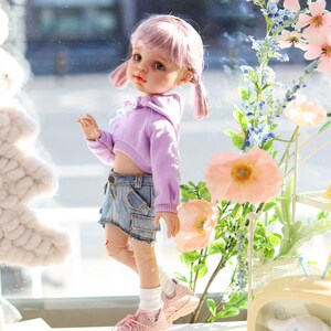 Pode incluir: Uma boneca rosa com cabelo rosa vestindo um top curto lavanda e shorts jeans. A boneca est&aacute; usando t&ecirc;nis rosa e meias brancas. A boneca est&aacute; em p&eacute; na frente de uma janela com flores e uma pequena televis&atilde;o amarela com um personagem de desenho animado branco dentro.