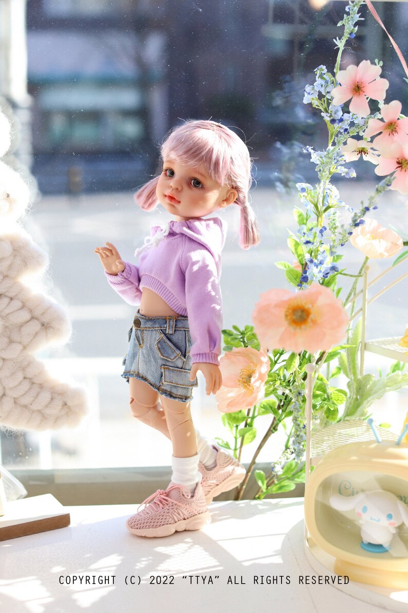 Pode incluir: Uma boneca rosa com cabelo rosa vestindo um top curto lavanda e shorts jeans. A boneca est&aacute; usando t&ecirc;nis rosa e meias brancas. A boneca est&aacute; em p&eacute; na frente de uma janela com flores e uma pequena televis&atilde;o amarela com um personagem de desenho animado branco dentro.