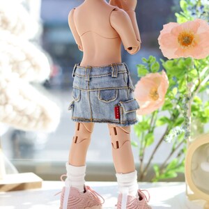 Pode incluir: Uma boneca vestindo uma saia de ganga azul claro com um logotipo vermelho, meias brancas e t&ecirc;nis rosa. A boneca tem cabelo rosa em rabo de cavalo e est&aacute; em p&eacute; em frente a uma janela.