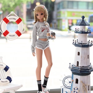 May include: A doll wearing a grey sweatshirt with the words "New York" on it, grey shorts, black and white striped socks, and pink and white sneakers. The doll is holding a small camera and is standing in front of a window with a life preserver and a lighthouse visible outside.