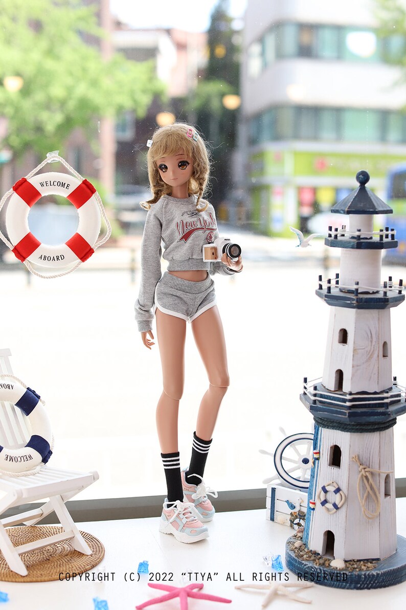 May include: A doll wearing a grey sweatshirt with the words "New York" on it, grey shorts, black and white striped socks, and pink and white sneakers. The doll is holding a small camera and is standing in front of a window with a life preserver and a lighthouse visible outside.