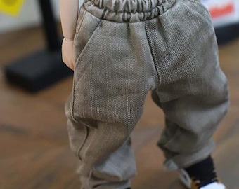 Puede incluir: Primer plano de un pantalón de muñeca gris con cintura elástica. El pantalón está hecho de un material similar al denim y tiene un corte holgado. La muñeca lleva zapatillas blancas, marrones y negras.