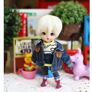 以下が含まれることがあります： 金髪の小さな人形が、青いデニムジャケット、青と白のストライプのシャツ、黄色と青のスニーカーを着用しています。人形は白い編み物の表面に立っています。