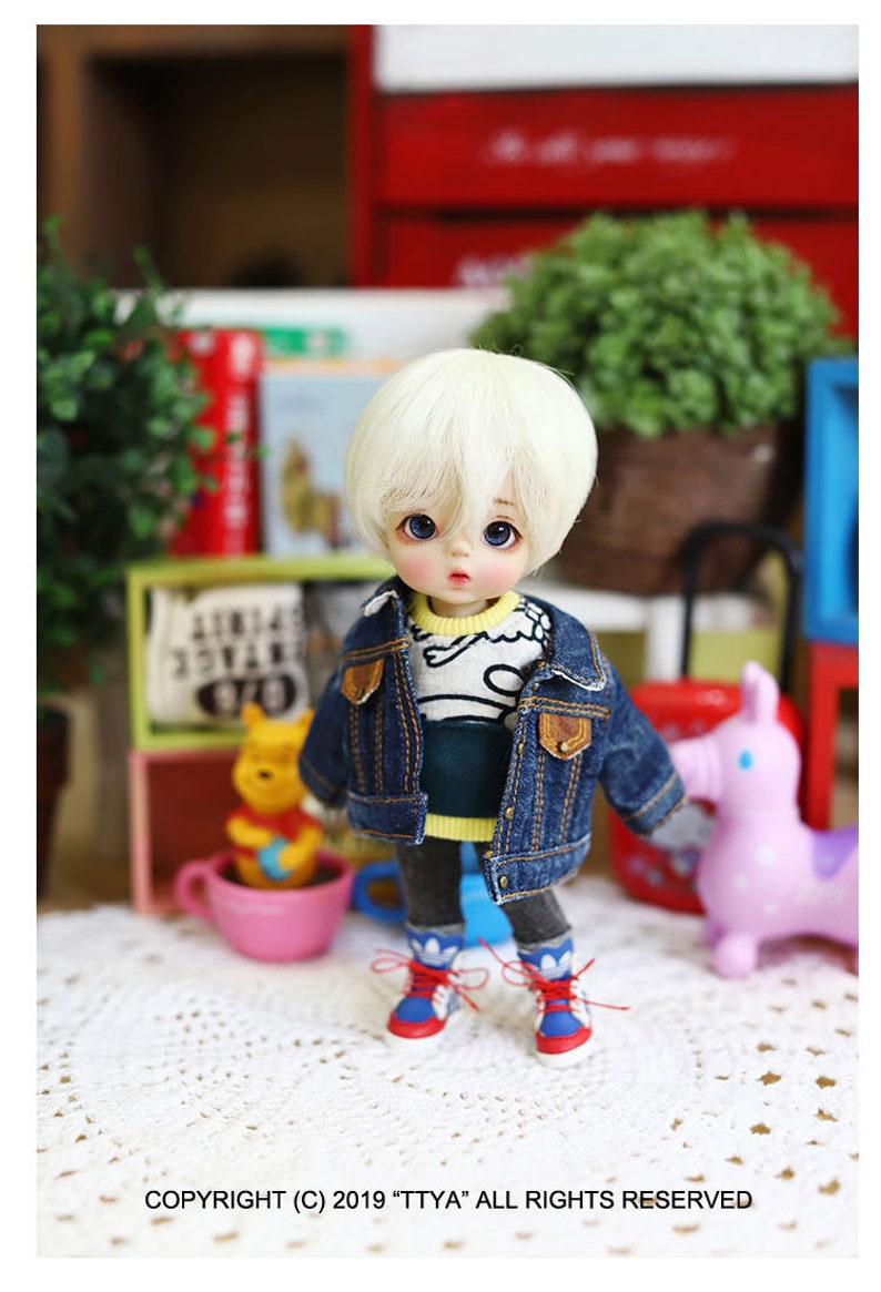 以下が含まれることがあります： 金髪の小さな人形が、青いデニムジャケット、青と白のストライプのシャツ、黄色と青のスニーカーを着用しています。人形は白い編み物の表面に立っています。