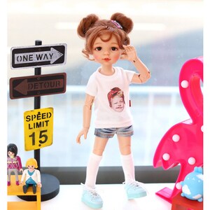 Puede incluir: Una mu&ntilde;eca con cabello casta&ntilde;o que lleva una camiseta blanca con una imagen impresa de una persona, pantalones cortos de mezclilla azul, calcetines blancos y zapatillas de deporte azul claro. La mu&ntilde;eca est&aacute; de pie sobre una mesa con un estante amarillo y una l&aacute;mpara de flamenco rosa. Tambi&eacute;n hay se&ntilde;ales de tr&aacute;fico que dicen "One Way" y "Detour" y una se&ntilde;al amarilla que dice "Speed Limit 15".