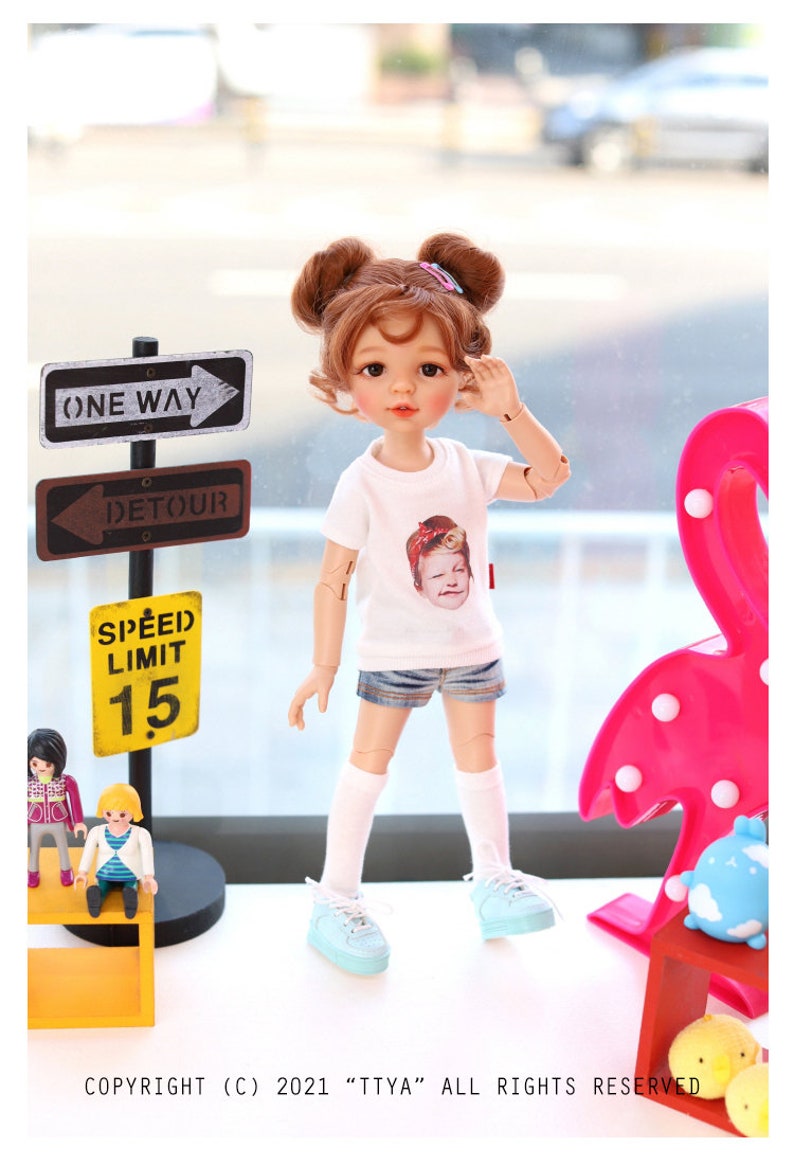 Puede incluir: Una mu&ntilde;eca con cabello casta&ntilde;o que lleva una camiseta blanca con una imagen impresa de una persona, pantalones cortos de mezclilla azul, calcetines blancos y zapatillas de deporte azul claro. La mu&ntilde;eca est&aacute; de pie sobre una mesa con un estante amarillo y una l&aacute;mpara de flamenco rosa. Tambi&eacute;n hay se&ntilde;ales de tr&aacute;fico que dicen "One Way" y "Detour" y una se&ntilde;al amarilla que dice "Speed Limit 15".