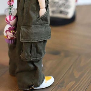 May include: Close-up of olive green cargo pants with a pocket detail. The pants are paired with white and yellow sneakers. A doll is wearing the pants. A decorative charm with a cupcake and candy is visible.