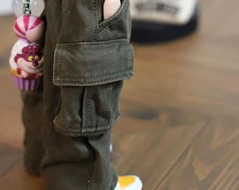 Puede incluir: Primer plano de pantalones cargo verde oliva con un detalle de bolsillo. Los pantalones se combinan con zapatillas blancas y amarillas. Una muñeca lleva los pantalones. Un charm decorativo con una magdalena y caramelos es visible.