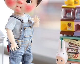May include: A small doll with short brown hair wearing a blue and white striped shirt and blue denim overalls. The doll has rosy cheeks and is holding its hand up in a wave. The doll is standing on a white surface.