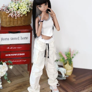 May include: A white doll with long black hair wearing a white tank top and white cargo pants with a black drawstring. The doll is standing on a wooden floor with a red dresser and a basket of flowers in the background.