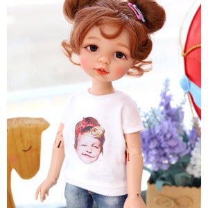 Puede incluir: Una mu&ntilde;eca con el pelo casta&ntilde;o peinado en dos mo&ntilde;os con pinzas para el pelo rosas. La mu&ntilde;eca lleva una camiseta blanca con una imagen impresa de la cara de una ni&ntilde;a, pantalones vaqueros azules y tiene ojos marrones. La mu&ntilde;eca est&aacute; de pie delante de un caballo de juguete de madera y un ramo de flores moradas.