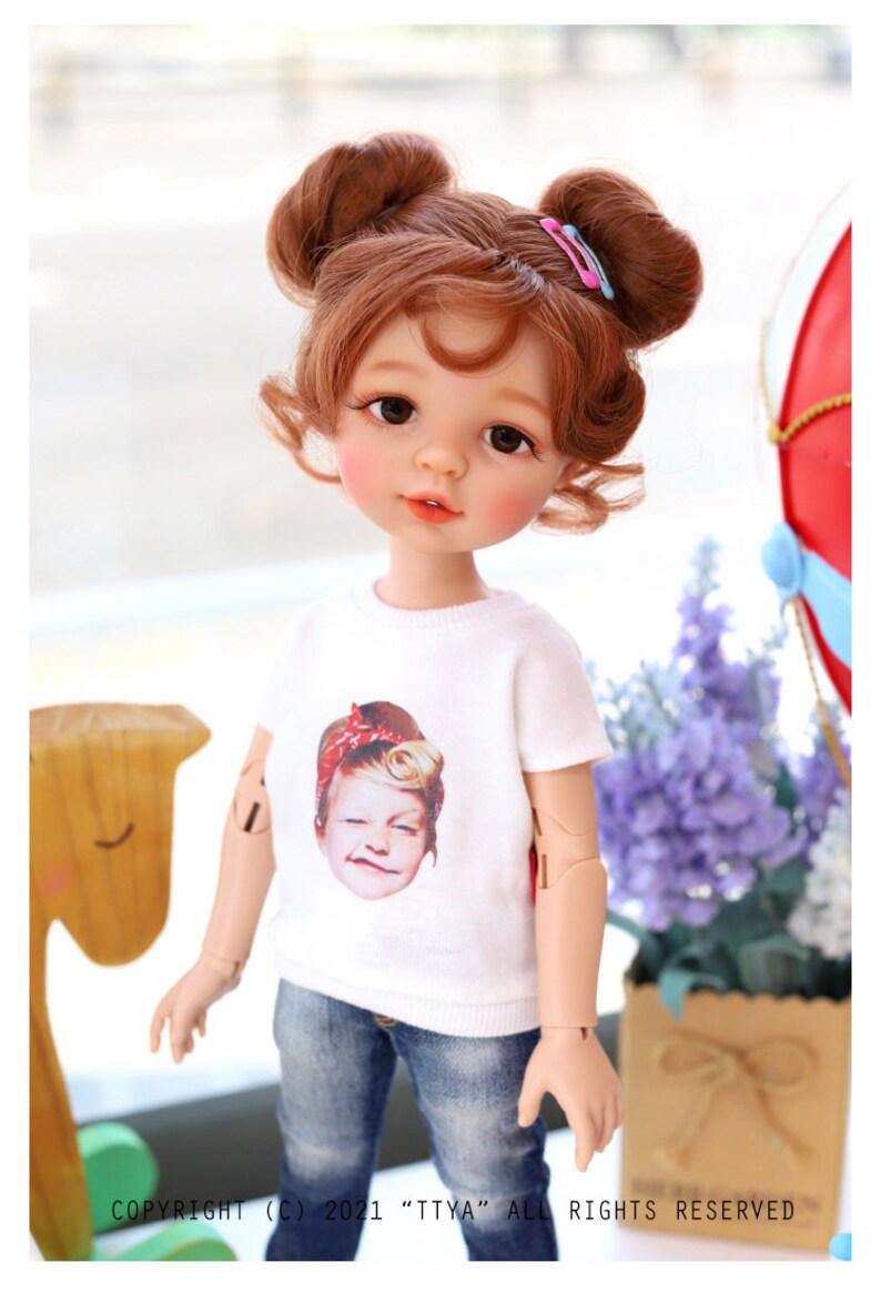 Puede incluir: Una mu&ntilde;eca con el pelo casta&ntilde;o peinado en dos mo&ntilde;os con pinzas para el pelo rosas. La mu&ntilde;eca lleva una camiseta blanca con una imagen impresa de la cara de una ni&ntilde;a, pantalones vaqueros azules y tiene ojos marrones. La mu&ntilde;eca est&aacute; de pie delante de un caballo de juguete de madera y un ramo de flores moradas.