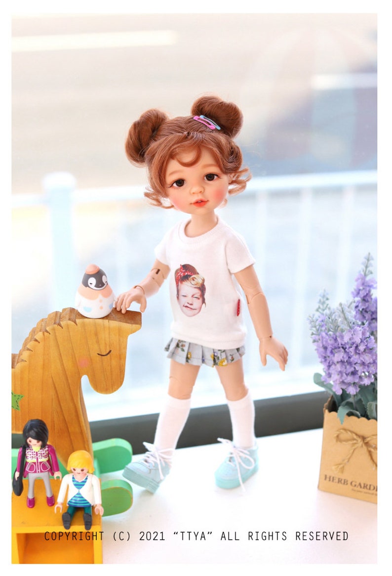 Puede incluir: Una mu&ntilde;eca con el pelo casta&ntilde;o en coletas que lleva una camiseta blanca con una imagen impresa de una ni&ntilde;a, una falda azul, calcetines blancos y zapatillas azules. La mu&ntilde;eca est&aacute; de pie junto a un caballo de balanc&iacute;n de madera y una peque&ntilde;a planta en maceta.