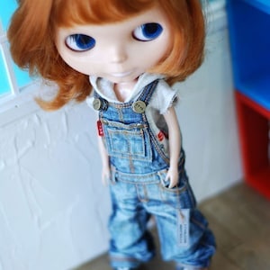 May include: A red-haired doll wearing a white shirt and blue denim overalls with a brown belt and metal buckles. The doll is standing on a wooden floor.