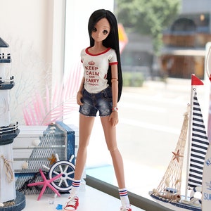 Puede incluir: Una mu&ntilde;eca con el pelo largo y negro que lleva una camiseta blanca con ribetes rojos y el texto "Keep Calm and Carry Myadoll" en rojo. La mu&ntilde;eca lleva pantalones cortos de mezclilla azul, calcetines a rayas rojas y blancas y zapatillas rojas. La mu&ntilde;eca est&aacute; de pie frente a una ventana con vista a una calle de la ciudad. Hay decoraciones n&aacute;uticas en una mesa frente a la mu&ntilde;eca.