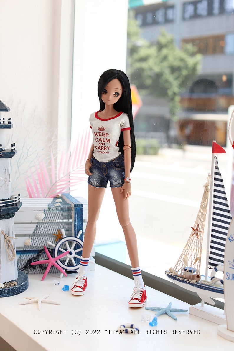 Puede incluir: Una mu&ntilde;eca con el pelo largo y negro que lleva una camiseta blanca con ribetes rojos y el texto "Keep Calm and Carry Myadoll" en rojo. La mu&ntilde;eca lleva pantalones cortos de mezclilla azul, calcetines a rayas rojas y blancas y zapatillas rojas. La mu&ntilde;eca est&aacute; de pie frente a una ventana con vista a una calle de la ciudad. Hay decoraciones n&aacute;uticas en una mesa frente a la mu&ntilde;eca.