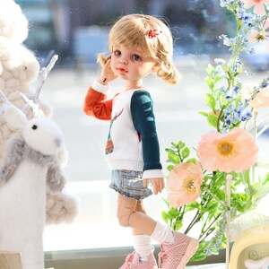 Pode incluir: Uma boneca com cabelo loiro vestindo um su&eacute;ter branco, laranja e turquesa com um desenho de veado, shorts jeans azul, meias brancas e t&ecirc;nis rosa. A boneca est&aacute; em p&eacute; em frente a uma janela com um animal de pel&uacute;cia branco e um buqu&ecirc; de flores rosas.