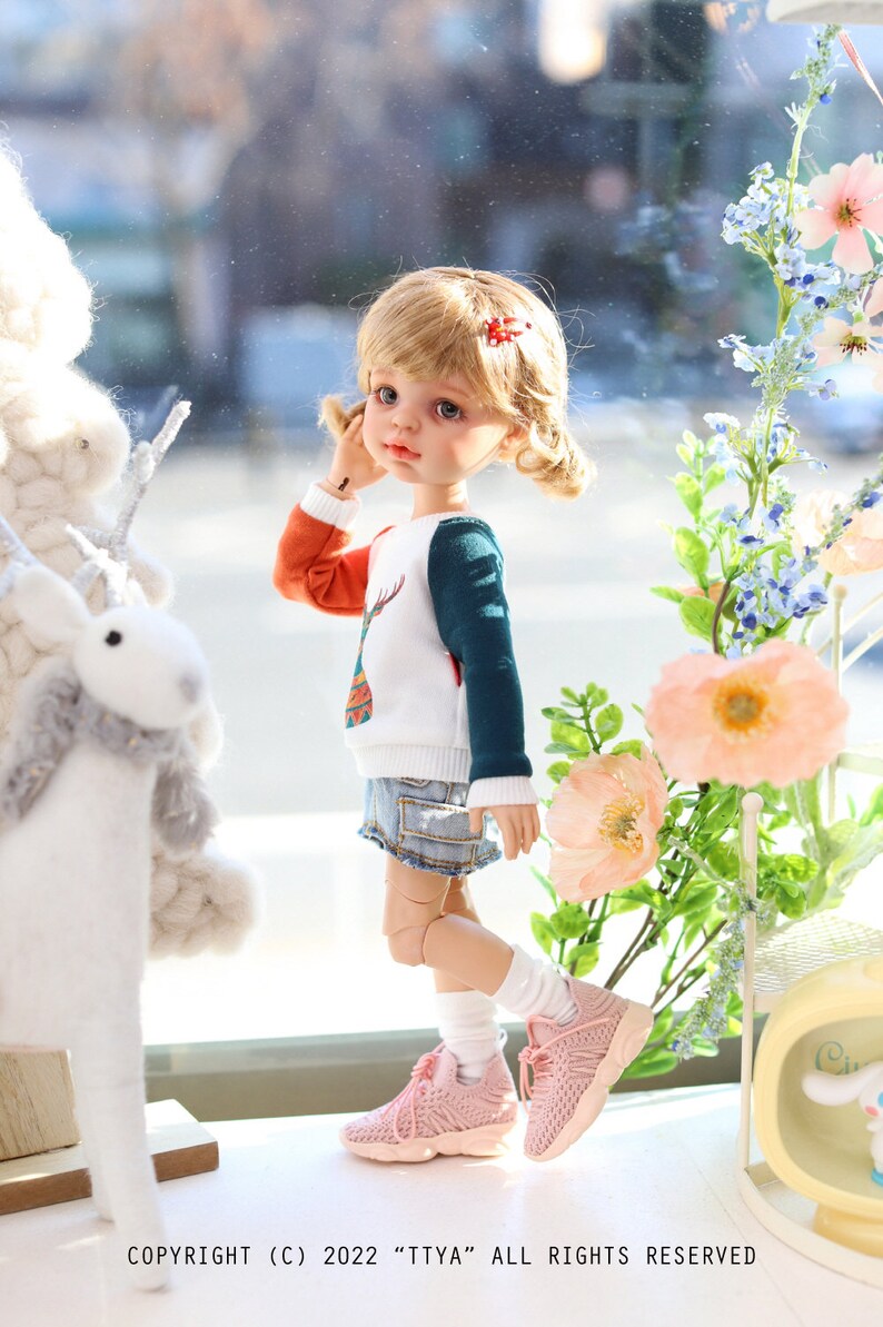 Pode incluir: Uma boneca com cabelo loiro vestindo um su&eacute;ter branco, laranja e turquesa com um desenho de veado, shorts jeans azul, meias brancas e t&ecirc;nis rosa. A boneca est&aacute; em p&eacute; em frente a uma janela com um animal de pel&uacute;cia branco e um buqu&ecirc; de flores rosas.