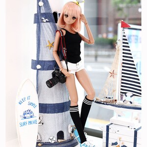 Puede incluir: Una mu&ntilde;eca con cabello rosado que lleva un top negro sin mangas, pantalones cortos blancos, calcetines negros hasta la rodilla con rayas blancas y zapatillas azules. La mu&ntilde;eca sostiene una c&aacute;mara negra y se apoya en un faro azul y blanco. La mu&ntilde;eca est&aacute; de pie sobre una superficie blanca con una tabla de surf blanca con el texto "Keep it Simple Surf More" en ella.