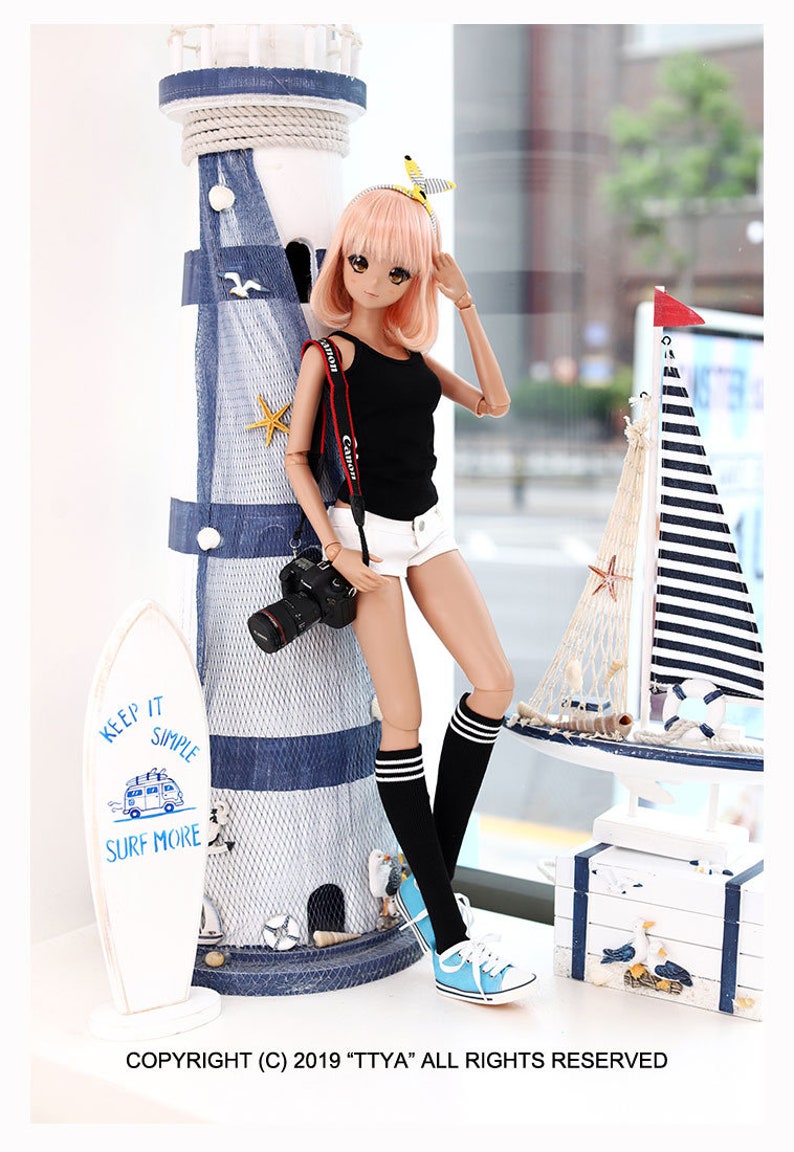 Puede incluir: Una mu&ntilde;eca con cabello rosado que lleva un top negro sin mangas, pantalones cortos blancos, calcetines negros hasta la rodilla con rayas blancas y zapatillas azules. La mu&ntilde;eca sostiene una c&aacute;mara negra y se apoya en un faro azul y blanco. La mu&ntilde;eca est&aacute; de pie sobre una superficie blanca con una tabla de surf blanca con el texto "Keep it Simple Surf More" en ella.