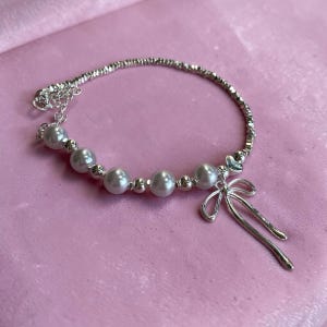 Puede incluir: Una pulsera plateada con perlas de imitación y un dije de lazo. La pulsera tiene una banda texturizada. La joya se exhibe sobre un fondo rosa, creando una estética femenina.