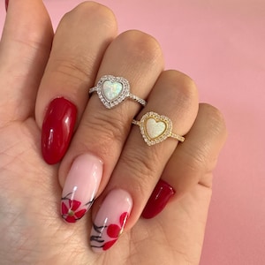 May include: Two silver rings featuring a heart-shaped opal stone set with small clear stones. The rings are worn on a hand with red and pink nail polish.