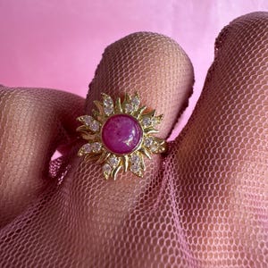 Puede incluir: Un anillo dorado con una gema rosa redonda y grande en el centro, rodeada por un diseño de sol con gemas claras más pequeñas. El anillo está sobre un fondo rosa.