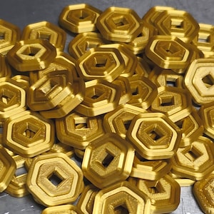 Puede incluir: Un montón de objetos geométricos dorados, cada uno con una forma exterior hexagonal y un agujero central cuadrado. Los objetos tienen un diseño en capas y parecen ser de metal. Están dispersos sobre una superficie.
