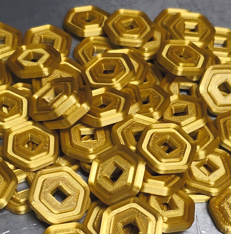 May include: A pile of gold-colored, geometric objects with a hexagonal outer shape and a square center hole. The objects have a layered design and appear to be made of a metallic material. The objects are scattered across a surface.