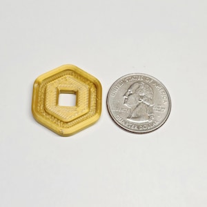 May include: A 3D-printed, gold-colored, hexagonal shape with a square center, next to a U.S. quarter. The object has a layered design and appears to be a token or a decorative item. The quarter is included for size comparison.