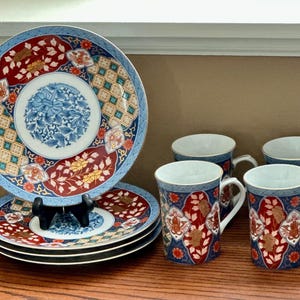 Op de afbeelding: Een set decoratieve porseleinen borden en mokken. De borden hebben een blauw, rood en goud bloemmotief. De mokken hebben een vergelijkbaar ontwerp. De borden zijn gestapeld op een houten oppervlak.