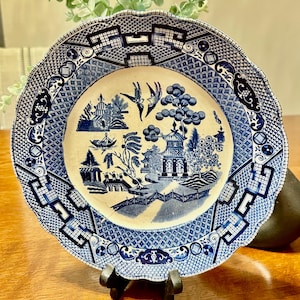 May include: A blue and white porcelain plate with a willow pattern design. The plate has a scalloped edge and a blue and white geometric pattern around the rim.