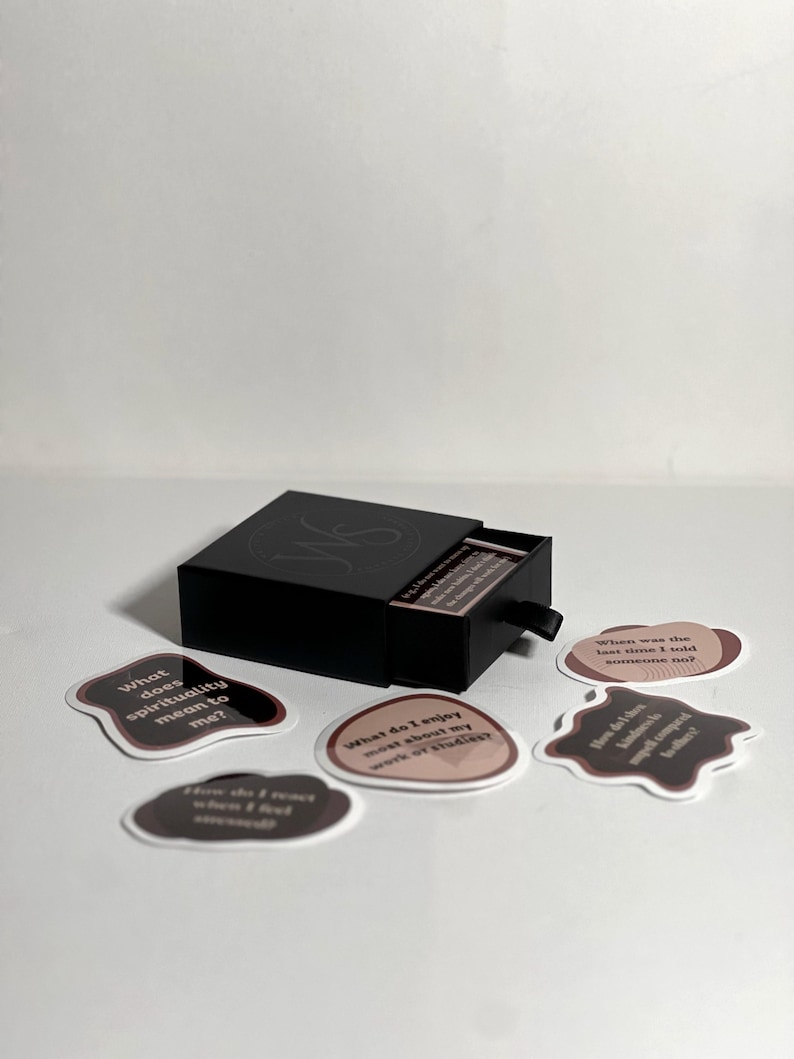 May include: A black box with a drawer open, revealing a stack of cards. The cards have brown and white text on a pink background. The text on the cards includes questions like "What does spirituality mean to me?" and "When was the last time I told someone no?"