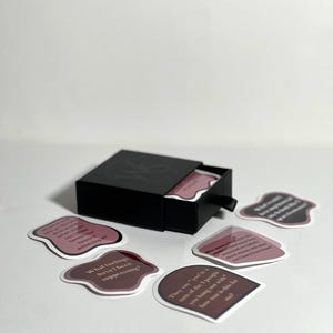 May include: A black box with a drawer open, revealing a set of pink and white cards with text. The cards have rounded edges and are scattered on a white surface. The text on the cards includes phrases like "What feelings have I been suppressing?" and "They say you're one of the 5 people you hang out with".