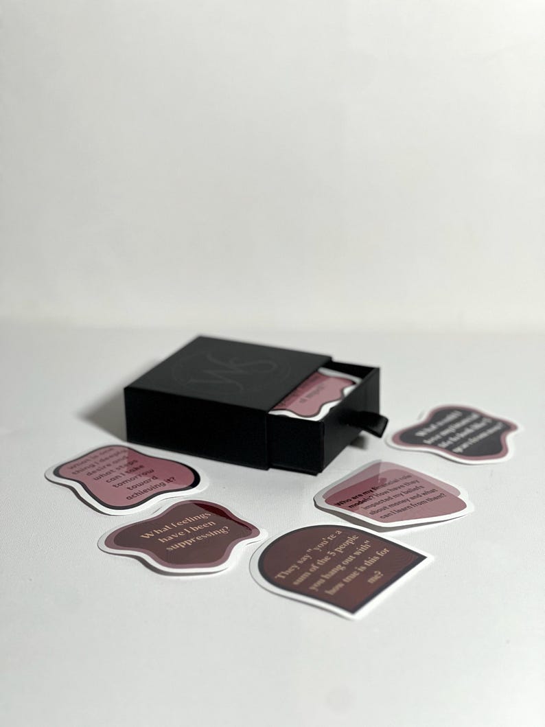 May include: A black box with a drawer open, revealing a set of pink and white cards with text. The cards have rounded edges and are scattered on a white surface. The text on the cards includes phrases like "What feelings have I been suppressing?" and "They say you're one of the 5 people you hang out with".