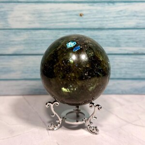 Puede incluir: Una esfera de labradorita verde oscuro con destellos iridiscentes azules y verdes, expuesta en un soporte de metal plateado.