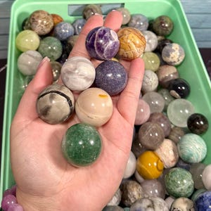 Può includere: Una mano tiene in mano una collezione di sfere di pietre preziose lucide in vari colori e motivi, tra cui ametista viola, avventurina verde e agata a strisce. Le sfere sono esposte in un vassoio di plastica verde, mostrando una varietà di trame di pietra naturale.