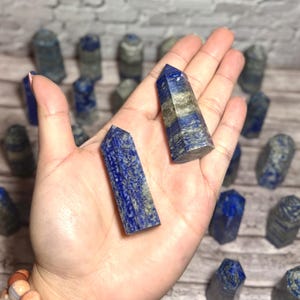 May include: Two blue lapis lazuli crystal towers are held in a hand, displaying their deep blue hue with gold inclusions. More crystal towers are visible in the background. The crystals are polished and faceted.