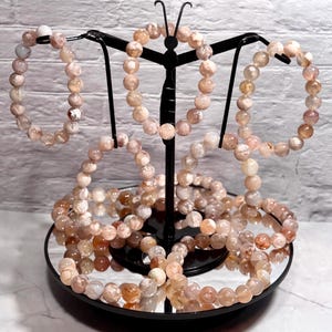 Puede incluir: Exhibición de varias pulseras de cuentas rosas y blancas. Las pulseras están hechas de piedras redondas y pulidas y están dispuestas sobre un soporte de metal negro con forma de mariposa y base de espejo. El fondo es una pared de ladrillos blancos.