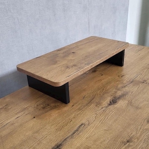 Puede incluir: Un soporte de monitor de madera con una superficie de vetas de madera marrón claro y patas negras. El soporte rectangular está elevado, proporcionando espacio debajo. La madera tiene un aspecto natural y texturizado.