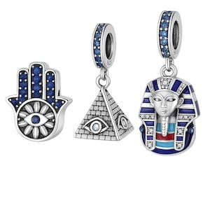 May include: Three silver charms with blue gemstone accents. One charm is a Hamsa hand with an eye design. Another is a pyramid with an eye. The third is a pendant of a pharaoh's head with blue and red details.