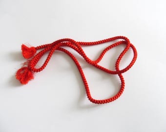 May include: A vibrant red cord with two tassels, lying on a white surface. The cord is tightly woven, and the tassels are made of a similar red, fringed material. The overall design is simple and elegant.